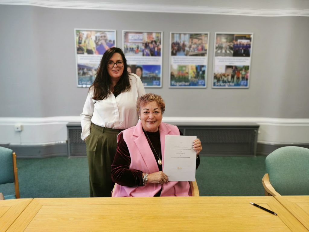 Photo of a women smiling sat by a text holding a document. A second woman stood behind smiling. Background shows a wall collage of photographs of Bury.