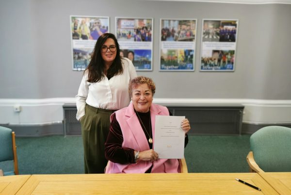 Photo of a women smiling sat by a text holding a document. A second woman stood behind smiling. Background shows a wall collage of photographs of Bury.